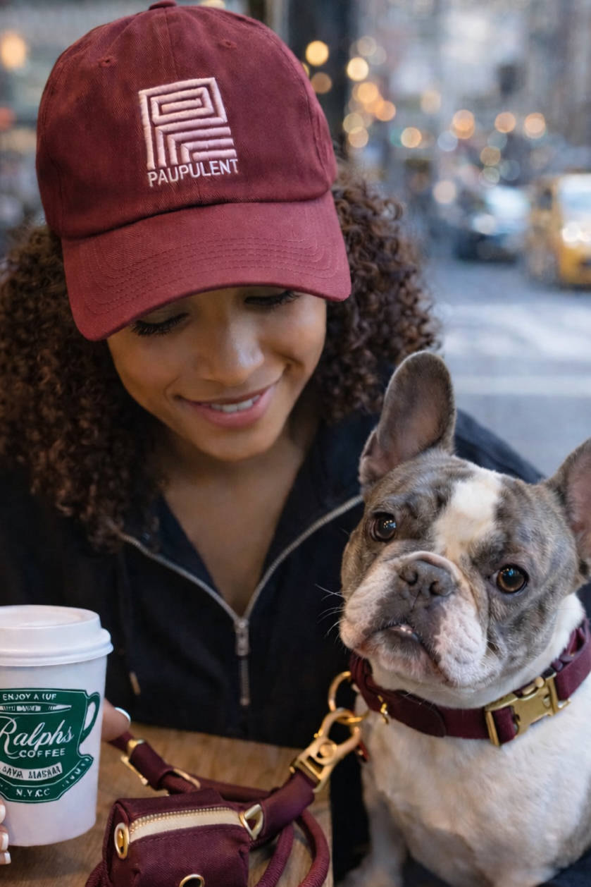Woman wearing a red cap with 'Palpulent' logo, holding a coffee cup, and sitting with a small dog on a city street.